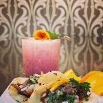 Food photo of pink cocktail with duck a la orange tacos on a plate. background is decorative wallpaper.