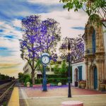 The historic train station with a large clock in the middle. Train tracks are on the left and a large jacaranda tree blooms in the backgroud.