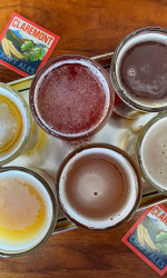 An overhead pov of a beer flight of 6 small glasses of beer on a tray with two business cards next to it that say Claremont Craft Beer
