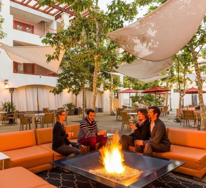 Four people sitting around a fire in a tree-filled courtyard of a hotel in Claremont for their spring break bucket list. 