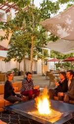 Four people sitting around a fire in a tree-filled courtyard of a hotel in Claremont for their spring break bucket list.