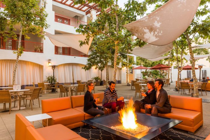 Four people sitting by a fire pit outside in a courtyard for happy hour.