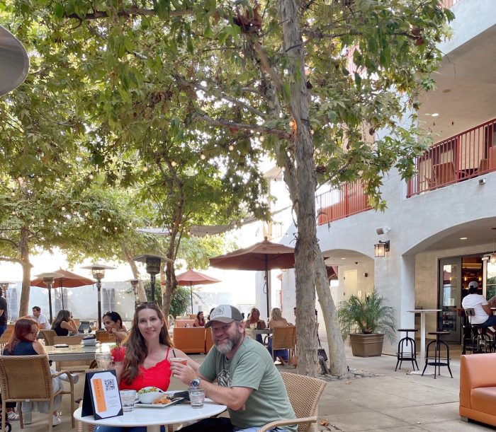 Man and woman sitting at table eating and drinking. Hotel courtyard in the background. 