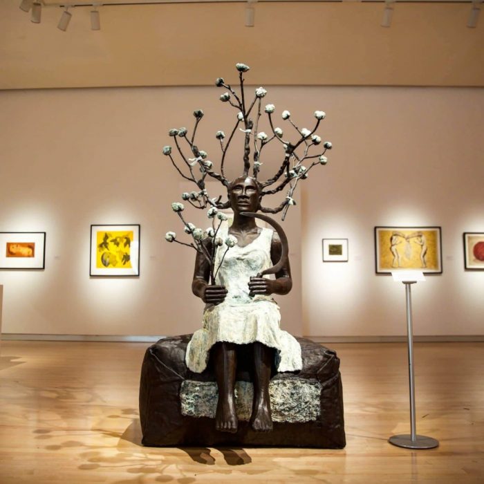 Bronze sculpture of a woman in white dress with tree growing out from behind her in gallery at a museum in Claremont