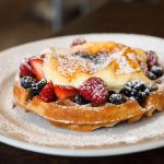 A waffle covered with strawberries, blackberries, creme brulee and powdered sugar.